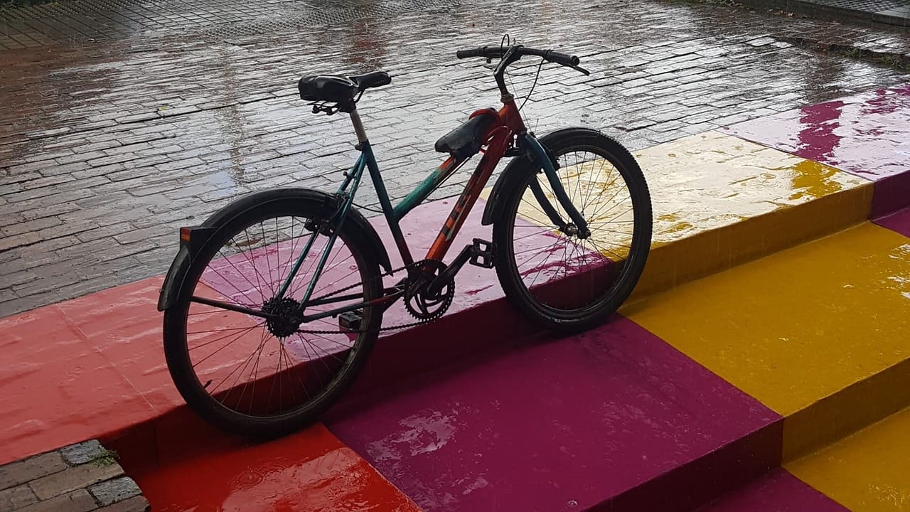 Bicycle rests against brick stairs. The stairs are playfully painted two shades of magenta and yellow. The brick is wet with rain.