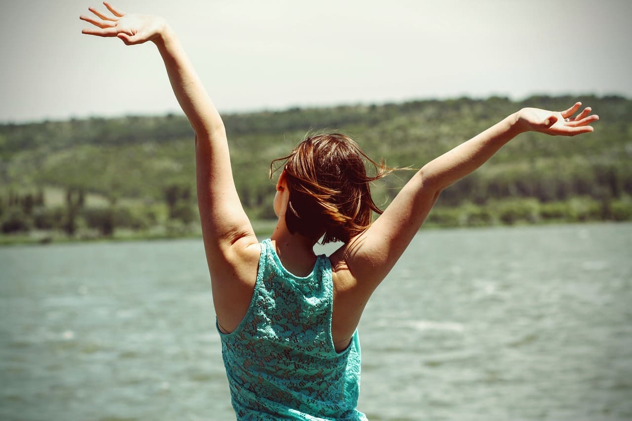 joyful person with long brown hair tied back, wearing a blue tank top, tossing hands into the air, stretching by the side of 